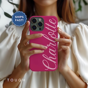 May include: A magenta phone case with the name "Charlotte" in a cursive font. The case is held in front of a person. The word "TOUGH" is at the bottom of the case. A blue and white sticker that says "SHIPS FAST!" is in the background.