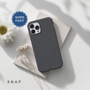 May include: A dark grey silicone phone case for an iPhone with a camera lens cutout. The case is on a white surface with a small white card that says "SNAP" on it.