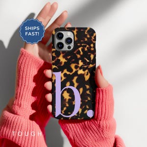 May include: A phone case featuring a tortoiseshell design and a lavender letter "b". The case is held in front of a pink sweater. The words "TOUGH" and "SHIPS FAST!" are also visible.