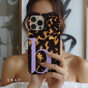 May include: A phone case featuring a tortoiseshell design and a large lavender letter "b". The case has a glossy finish and a camera aperture. The word "SNAP" is printed in white at the bottom.