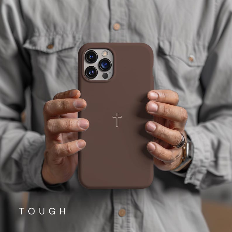 May include: A brown phone case with a cross symbol. The case has a matte finish and is held in front of a person. The word "TOUGH" is printed at the bottom of the image.