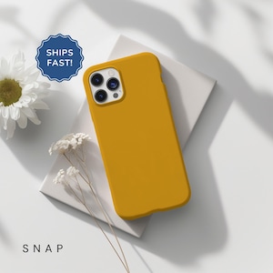 May include: Mustard yellow phone case with a sleek design. The case is resting on a white surface, with a white flower and delicate sprigs nearby. The text "SNAP" is at the bottom of the image.