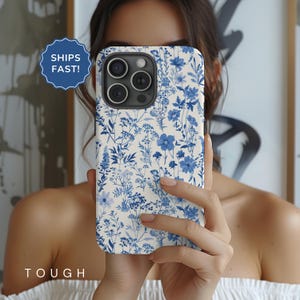 May include: A white phone case with a blue floral pattern. The case has a camera lens cutout and a cutout for the phone's buttons. The case is being held by a person's hand.