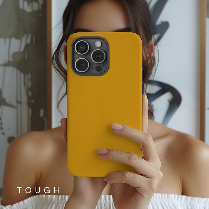 May include: Mustard yellow phone case with a black camera lens frame. The word "TOUGH" is printed in white at the bottom of the image. The phone case is shown in a close-up view.