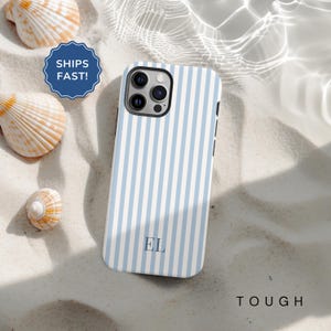 May include: A smartphone case featuring a blue and white striped design, with the initials "EL" in the center. The case is displayed on a sandy surface, alongside seashells. The word "TOUGH" is printed on the bottom right.