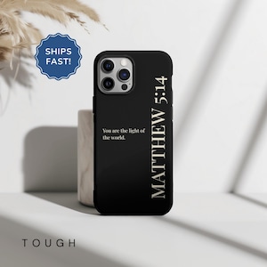 Op de afbeelding: Zwarte telefoonhoes met de tekst "You are the light of the world" en "MATTHEW 5:14" in gouden letters. De hoes wordt getoond op een wit oppervlak met een neutrale achtergrond. Het woord "TOUGH" staat onderaan afgedrukt.