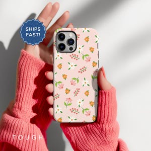 May include: A floral phone case with a pink background, featuring a pattern of white, orange, and pink flowers with green leaves. The case is held in a person's hands, wearing a pink sweater. A blue badge says "SHIPS FAST!" and the word "TOUGH" is visible.