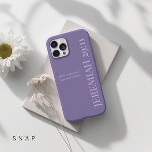 Pu&ograve; includere: Custodia per telefono lilla con la scritta "JEREMIAH 29:11" in verticale e "Plans to give you hope and a future." in orizzontale. La custodia &egrave; su una superficie bianca con un fiore bianco e rametti.