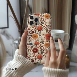 May include: A white iPhone case with a floral pattern featuring red, orange, and yellow flowers. The case has a camera lens cutout and a white background.