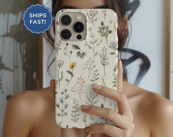 Può includere: Una custodia per telefono bianca con un motivo floreale che presenta varie foglie verdi, fiori rosa e gialli. La custodia ha un ritaglio per l'obiettivo della fotocamera.
