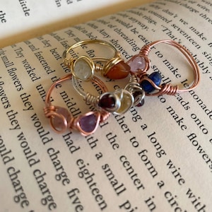 May include: A collection of handmade wire-wrapped rings with various gemstones, including amethyst, rose quartz, and carnelian. The rings are displayed on the open pages of a book, showcasing the jewelry's delicate craftsmanship and natural stone colors.