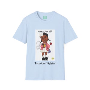 Könnte beinhalten: Hellblaues T-Shirt mit einer Cartoon-Illustration eines jungen Mädchens, das ein Buch und einen Apfel hält. Der Text "WHO AM I?" befindet sich über der Illustration und "Freedom Fighter!" darunter.