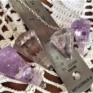 May include: Three amethyst crystal pendulums with silver chains. The pendulums are different sizes and shapes. The largest pendulum is about 2.5 inches long. The pendulums are on a white crocheted surface.