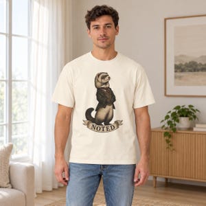 May include: A cream-colored short-sleeve t-shirt featuring a graphic illustration of a ferret dressed in a dark suit, vest, and monocle. Below the ferret is a beige ribbon banner with the word NOTED printed in bold, dark capital letters.