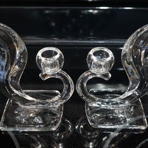 May include: A pair of clear glass candle holders with a unique, curved design. Each holder features a round, open top for a candle and a square base.