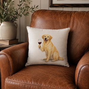 May include: A square throw pillow featuring a printed illustration of a yellow Labrador retriever sitting upright and holding a stemmed glass filled with red wine. The pillow has a light beige, textured fabric background and a simple square shape.