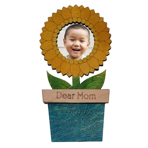 May include: Wooden photo frame shaped like a sunflower with a child's photo in the center. The sunflower is in a blue pot with the text "Dear Mom Thanks for helping me grow".