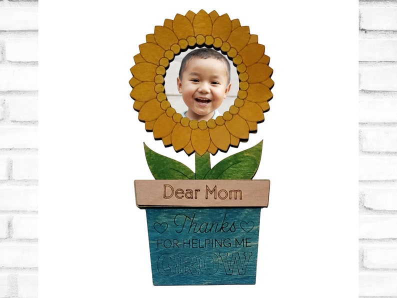 May include: Wooden photo frame shaped like a sunflower with a child's photo in the center. The sunflower is in a blue pot with the text "Dear Mom Thanks for helping me grow".