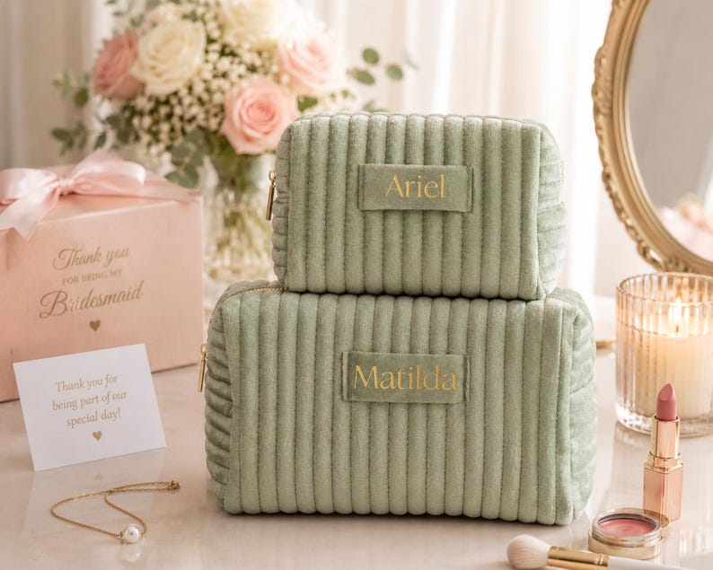 a set of two green makeup bags with gold text, placed on a table with a vase of flowers, a card, and other personal items.