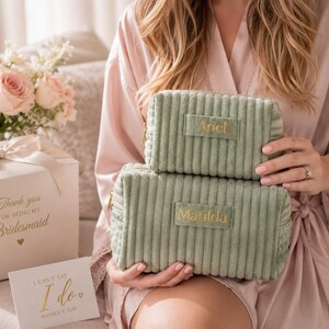 A woman in a pink robe holding a green makeup bag with the name "Ariel" on it, next to a gift box with a bow and a card that says "Thank you for being my Bridesmaid".