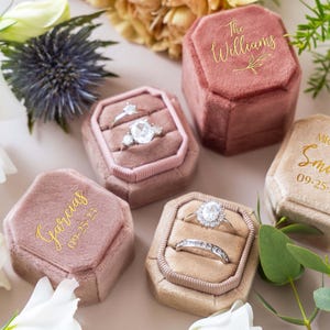 May include: A collection of velvet ring boxes in various shades of pink and beige. Each box is octagonal and features a gold inscription with names and dates. Several boxes contain diamond rings, showcasing a wedding theme.