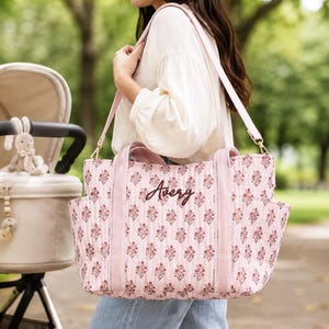 May include: A quilted tote bag in a soft pink hue featuring a repeating dark pink floral pattern and the name Avery embroidered in dark script on the front. The bag includes two sturdy pink fabric handles and a long adjustable shoulder strap.
