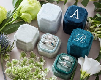 May include: A collection of hexagonal velvet ring boxes in shades of light blue, dark blue, and teal. Two boxes are open, revealing silver rings with clear stones. The dark blue and teal boxes feature the white embroidered letters A and R.