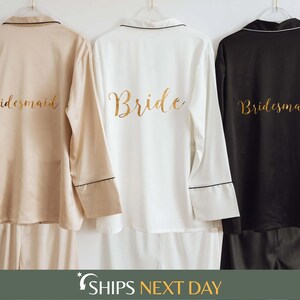 May include: Three long-sleeved pyjama shirts hanging on white hangers. The left shirt is beige, the centre is white, and the right is black. Each shirt features gold script text on the back, with the centre reading Bride and the others Bridesmaid.