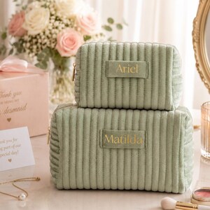 a set of two green makeup bags with gold text, placed on a table with a vase of flowers, a card, and other personal items.