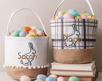 May include: Two Easter baskets filled with colorful eggs. One is white with a bunny design and the name "Sarah". The other is plaid with a bunny design and the name "Noah". Both have handles and burlap bottoms.