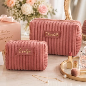 a pink makeup bag with the name "Evelyn" written on it, placed on a vanity with a mirror and other makeup items. There is also a small gift box with a bow and a vase of flowers in the background.