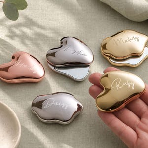 May include: Five heart-shaped compact mirrors with metallic finishes in rose gold, silver, and gold. Each mirror features a custom name engraved in script font: Hailey, Lydia, Melody, Daisy, and Ruby. The mirrors have a smooth, reflective surface.