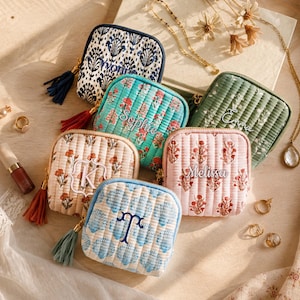 Op de afbeelding: Een collectie van zes vierkante, gewatteerde stoffen tasjes met diverse bloemen- en schelpenpatronen, elk voorzien van een ritssluiting, een decoratief kwastje en gepersonaliseerd borduurwerk met namen zoals Sophie, Emma, Melissa en monogrammen.