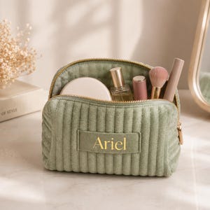 A green makeup bag with the name "Ariel" printed on it, containing various makeup items such as a compact, powder, and a brush.