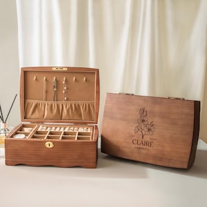 May include: Two wooden jewelry boxes with a medium brown finish. One box is open, revealing a tan velvet interior with necklace hooks, a pocket, and ring slots. The closed box features a laser-engraved floral design and the text EST 2024 CLAIRE CAMPBELL.