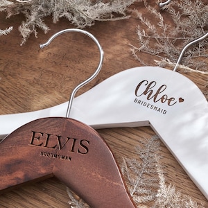 May include: Two wooden clothes hangers with silver metal hooks. One hanger is dark brown with the engraved text ELVIS and GROOMSMAN. The other hanger is white with the printed text Chloe and a heart symbol followed by the word BRIDESMAID.