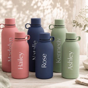 May include: Six matte-finish water bottles in pink, navy blue, and sage green. Each bottle features a screw-top lid with a side loop and is personalised with white vertical text reading Madelyn, Hailey, Natalie, Rose, Kennedy, and Ashley.