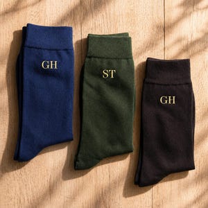 May include: Three pairs of socks in navy blue, forest green, and black, each with embroidered initials. The socks are displayed on a light wood surface. The navy sock has "GH", the green sock has "ST", and the black sock has "GH".