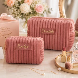 a pink makeup bag with the name "Evelyn" written on it, placed on a vanity with a mirror and other makeup items. There is also a small gift box with a bow and a vase of flowers in the background.