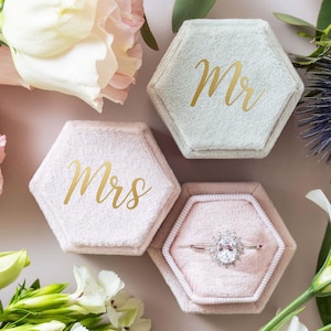 May include: Two hexagonal velvet ring boxes in light blue and soft pink. The light blue box lid features the gold text Mr, while the pink box lid features the gold text Mrs. The open pink box contains a silver ring with a central oval stone.