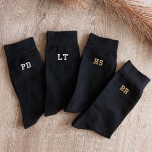 May include: Four black socks with ribbed cuffs, each featuring embroidered initials. Two socks display the letters PD and LT in white thread, while the other two socks feature the letters HS and BR embroidered in a metallic gold thread.