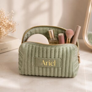 A green makeup bag with the name "Ariel" printed on it, containing various makeup items such as a compact, powder, and a brush.