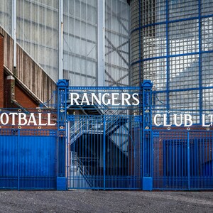 Könnte beinhalten: Ein blaues Metalltor mit der Aufschrift "Rangers Football Club Ltd." in weißen Buchstaben. Das Tor steht vor einem Backsteingebäude mit einer Glasfassade.