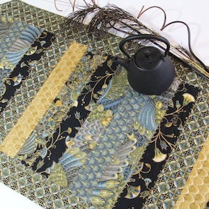 Puede incluir: Camino de mesa decorativo con un diseño de plumas de pavo real en tonos azules, dorados y negros. Una tetera negra y ramas decorativas encima. El camino de mesa mide aproximadamente 152 cm de largo.