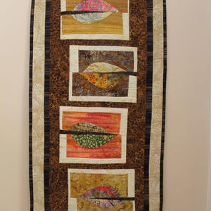 K&ouml;nnte beinhalten: Ein rechteckiger Wandbehang mit braunem Leopardenmuster-Hintergrund und vier rechteckigen Tafeln. Jede Tafel zeigt ein blattartiges Design mit verschiedenen Farben, darunter Rosa, Gr&uuml;n und Orange, und einen schwarzen horizontalen Streifen. Der Rand ist eine Mischung aus Creme, Braun und Schwarz.