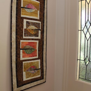K&ouml;nnte beinhalten: Ein rechteckiger Wandbehang mit einem braunen und beigefarbenen Musterhintergrund. Er zeigt f&uuml;nf rechteckige Tafeln mit abstrakten blattartigen Designs in Orange-, Rosa- und Gr&uuml;nt&ouml;nen. Das St&uuml;ck ist mit einem dunkelbraunen Rand und einem helleren beigefarbenen Innenrand versehen.