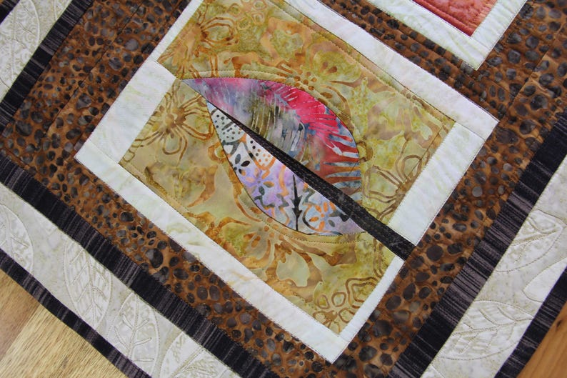 K&ouml;nnte beinhalten: Nahaufnahme eines handgefertigten Quilts mit einem Blattdesign. Der Quilt zeigt eine Kombination aus gemusterten Stoffen in Braun-, Beige- und Wei&szlig;t&ouml;nen. Das zentrale Design umfasst eine farbenfrohe Blattform mit rosa, lila und wei&szlig;en Akzenten.