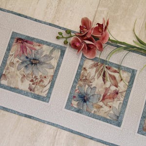 May include: A long rectangular table runner featuring a repeating pattern of square panels. Each panel contains a floral print with blue and pink flowers on a cream background, framed by a light blue border and a textured white fabric border.