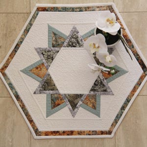 Peut inclure: Nappe hexagonale matelassée blanche avec un motif en étoile. L'étoile est composée de triangles dans les tons de gris, de sarcelle et d'orange. La bordure présente un motif moucheté dans des couleurs similaires. Une petite orchidée en pot avec des fleurs blanches est posée sur la nappe.