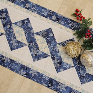 May include: A quilted table runner featuring a repeating zigzag pattern in navy blue and white fabric with snowflake motifs. The runner is decorated with a sprig of holly with red berries and two gold and silver textured decorative spheres.